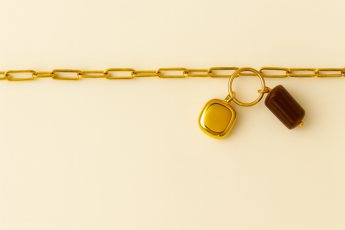 Collar dorado de eslabones con dos charms dorado y marrón sobre fondo crema editorial.